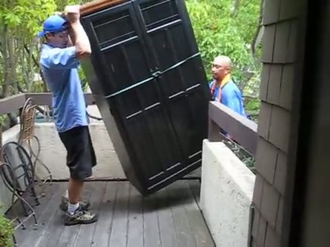 two movers carrying a tall black armoire across an outdoor deck, face visible