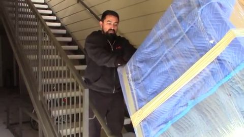 mover carrying a shrink-wrapped dresser through an apartment stairwell, face visible