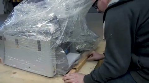 worker fitting a shrink-wrapped item onto a wooden crate base, face visible