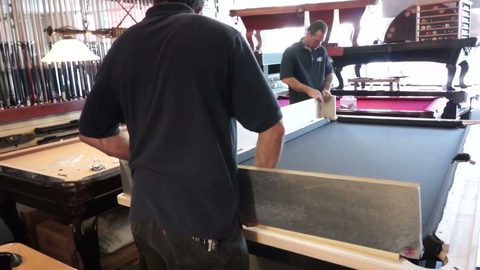 mover carrying a slate rail piece while crew works the pool table behind, face visible
