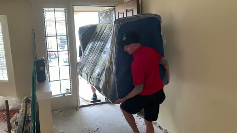 Two movers carrying a wrapped sofa through a front doorway, face visible