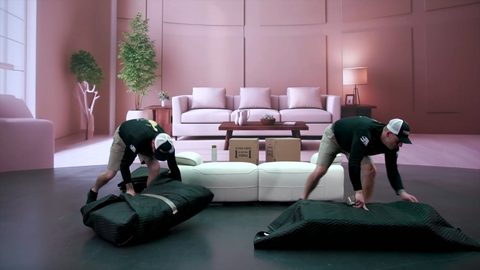 Two movers crouching to spread moving blankets on the floor beside a white reclining loveseat