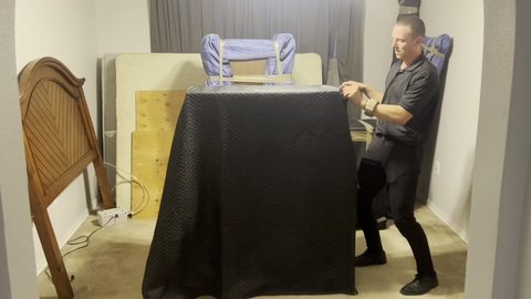 Mover wrapping a nightstand in a black moving blanket in a cleared bedroom, face visible